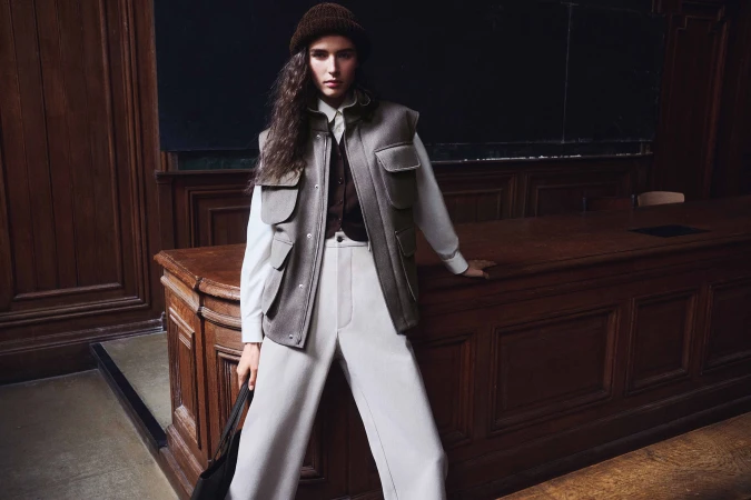 Model in a brown beanie, taupe utility vest, and white wide-leg trousers leaning against a wooden lecture desk in Paris.