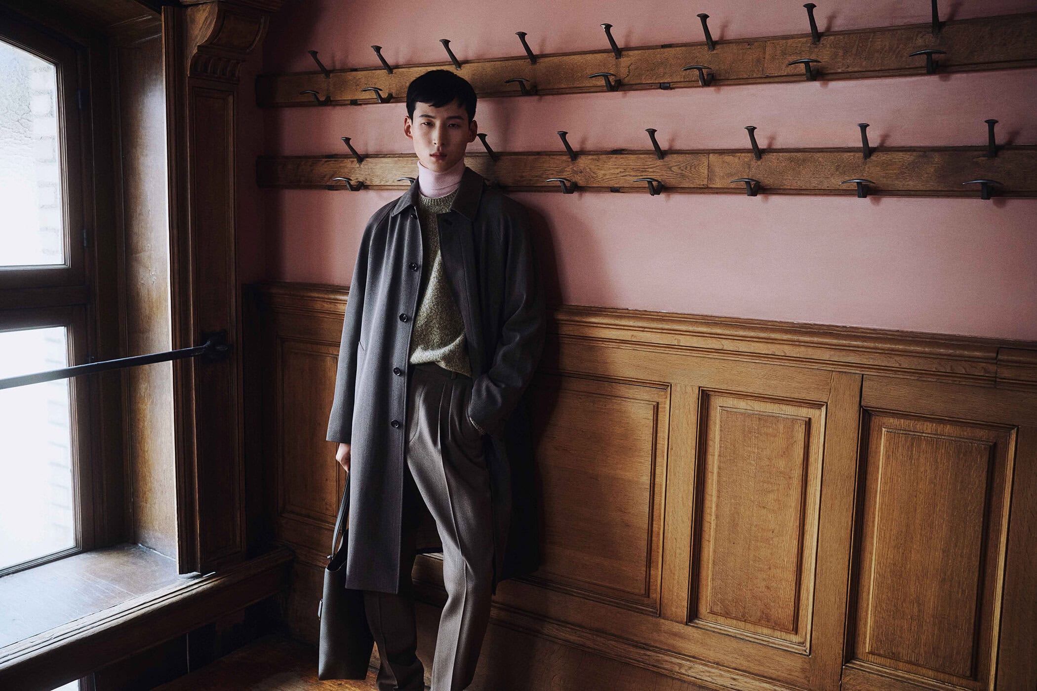 East Asian man in a charcoal overcoat, turtleneck, and pleated trousers leaning against a wood-paneled wall next to a window.