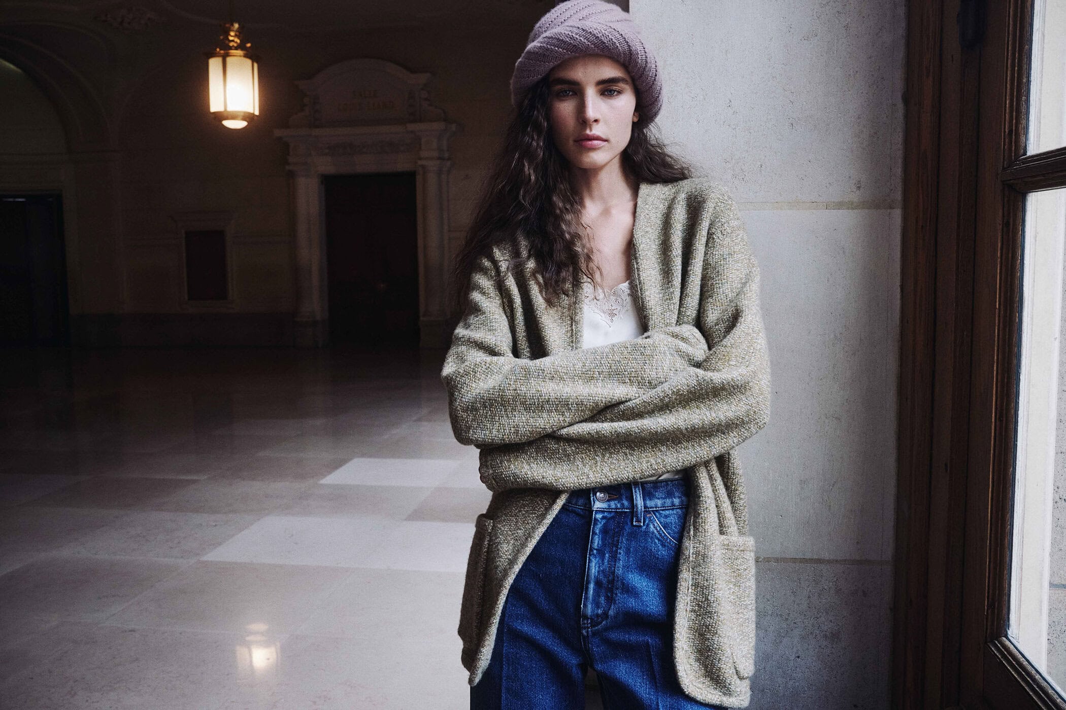 Libby Taverner posing in an oversized sage knit cardigan, blue jeans, and a pink knit hat against a stone wall.