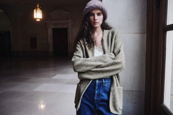 Libby Taverner posing in an oversized sage knit cardigan, blue jeans, and a pink knit hat against a stone wall.
