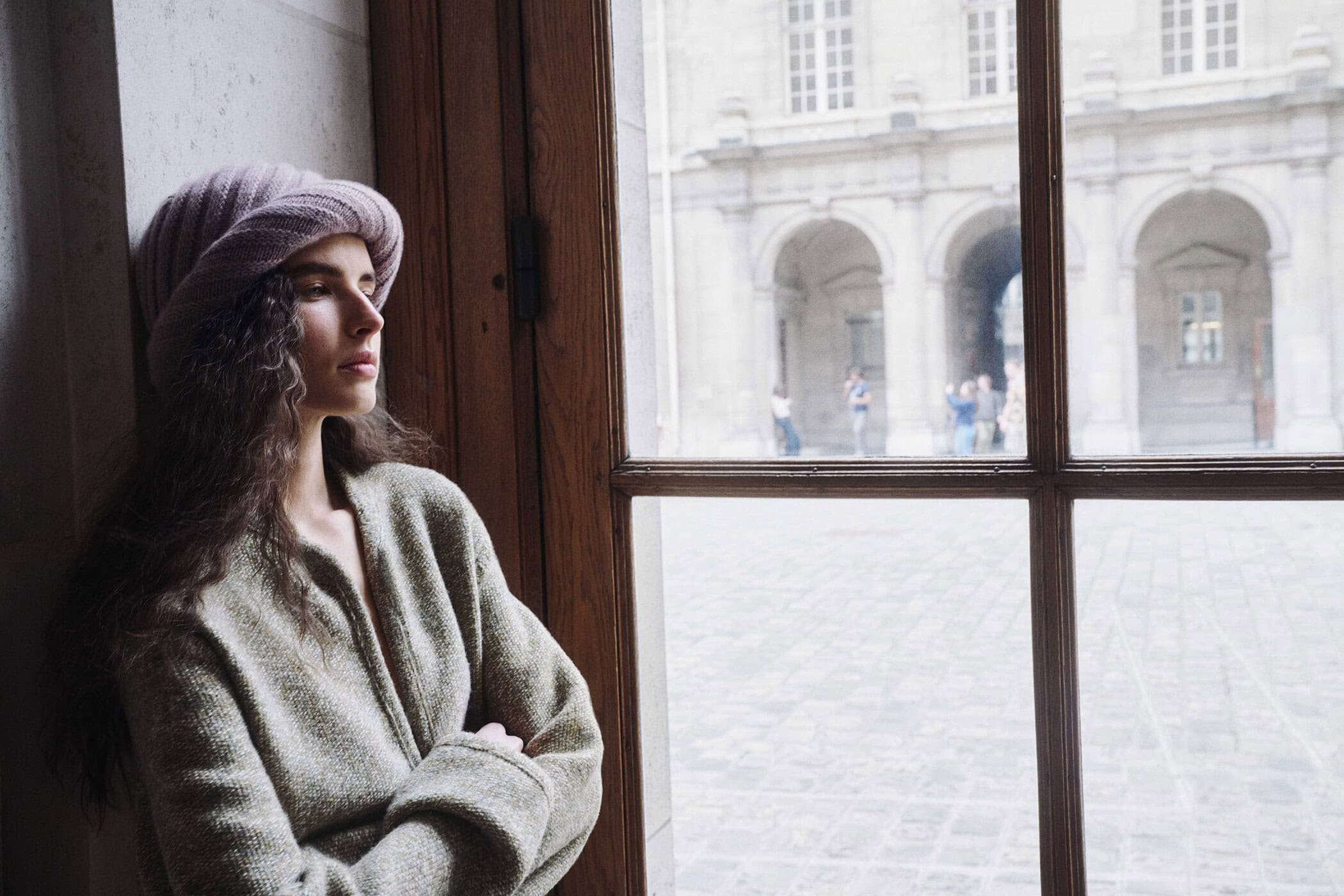 Model in a sage green knit cardigan and pink ribbed knit hat looking out a window overlooking a historic stone courtyard in Paris.