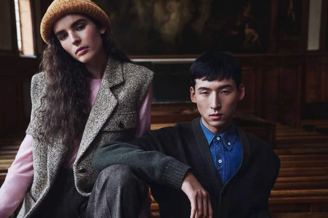 Two fashion models pose in layered wool knitwear and tailored tweed inside a wood-paneled hall in Paris.