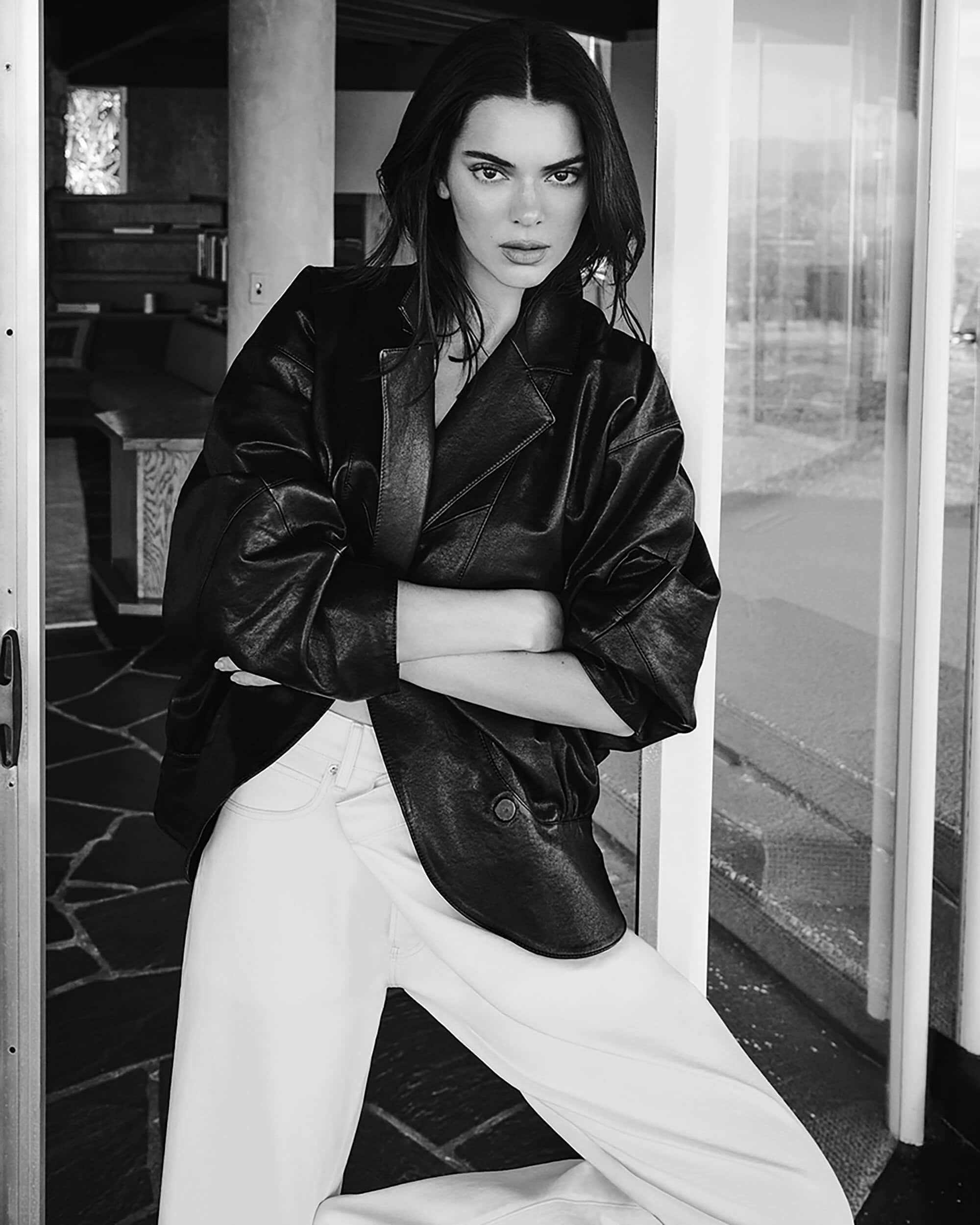 Kendall Jenner poses wearing an oversized black leather jacket and white wide-leg trousers leaning in an open doorway.