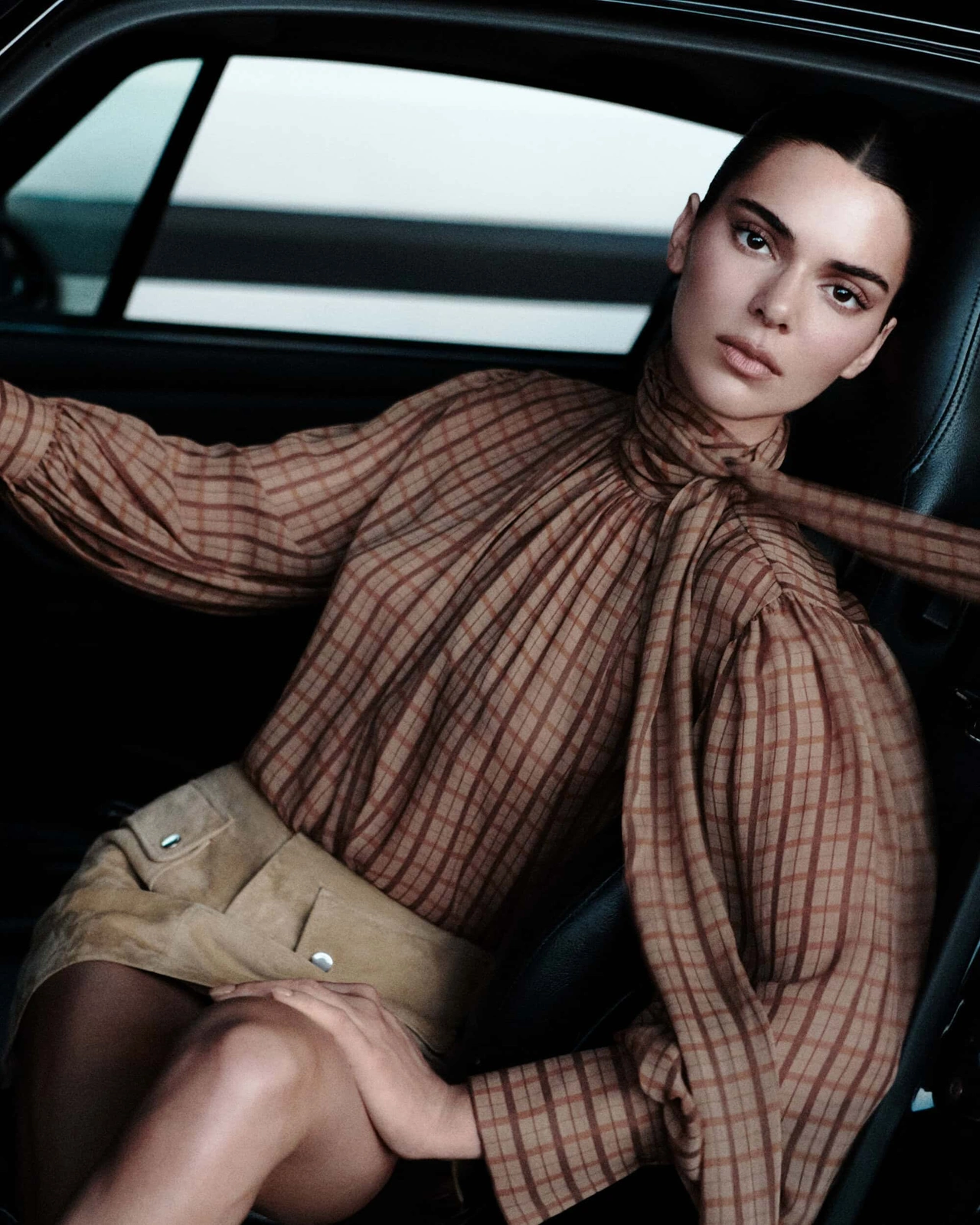 Kendall Jenner wearing a brown plaid high-neck blouse and tan suede mini skirt seated inside a car for FRAME.