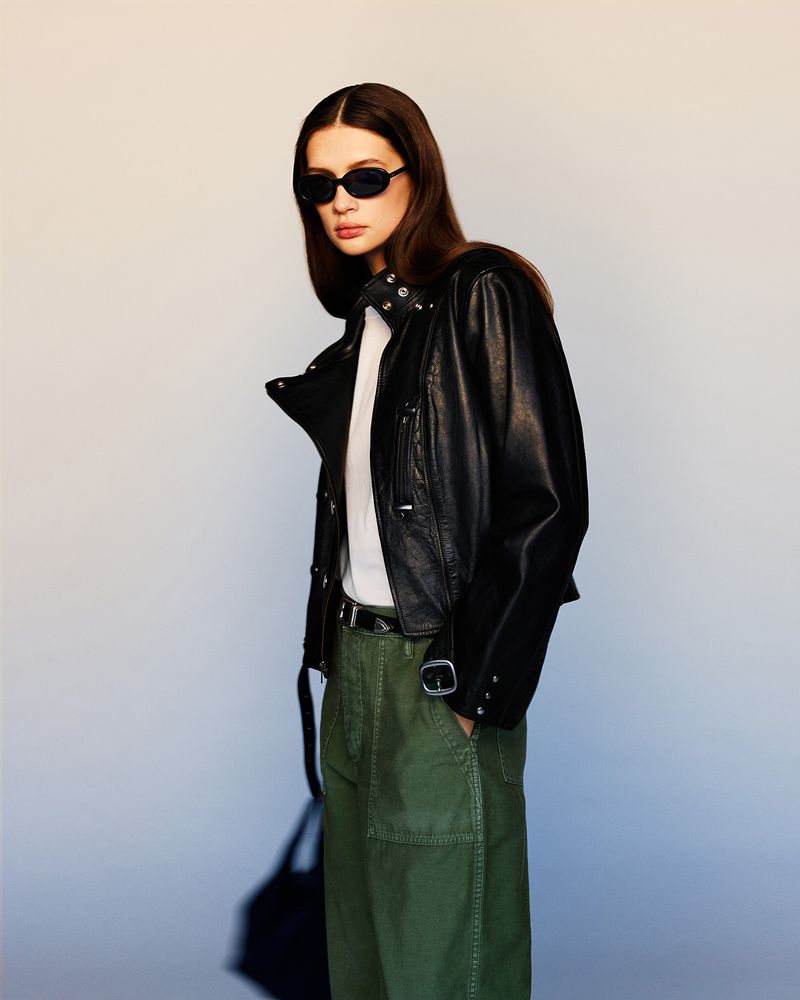 A woman poses in a studio, wearing a black leather jacket, white shirt, and green pants, accessorized with sunglasses and a belt.