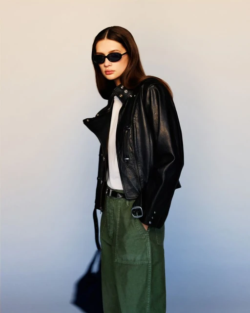 A woman poses in a studio, wearing a black leather jacket, white shirt, and green pants, accessorized with sunglasses and a belt.