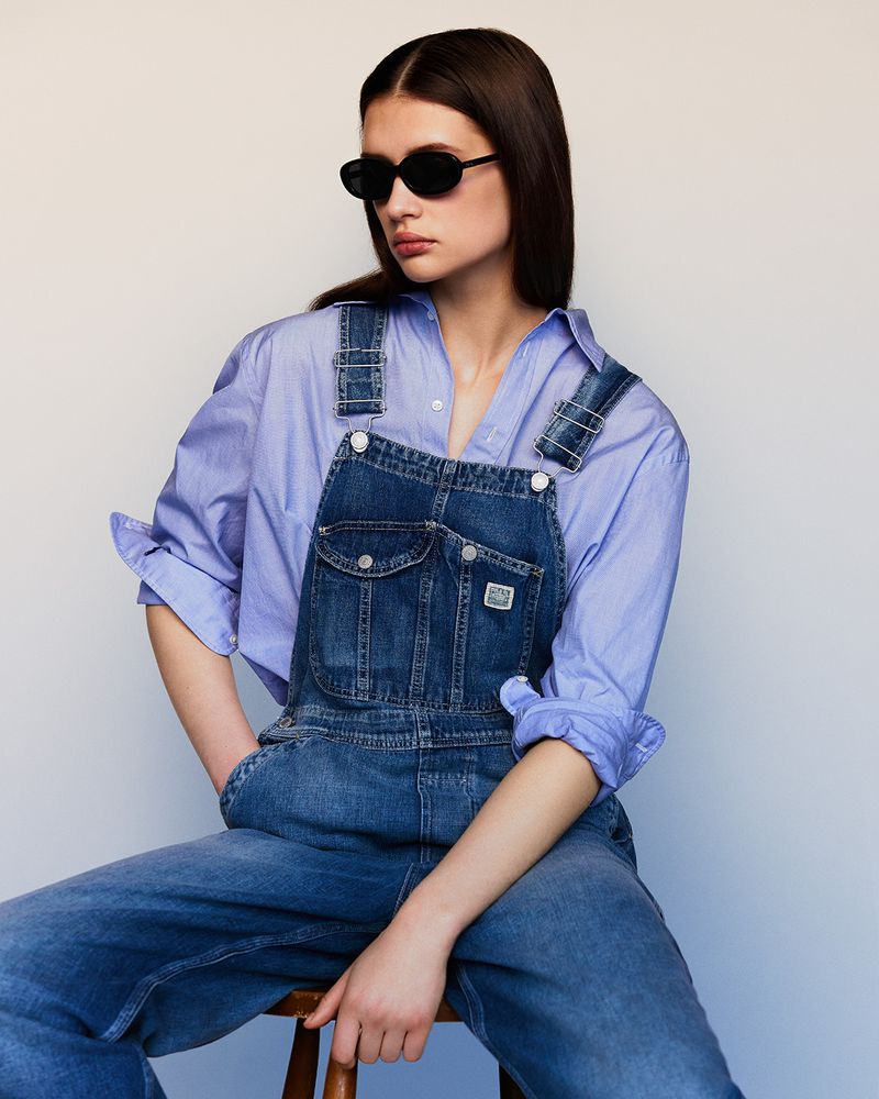 A woman in a blue denim overall and a light purple button-down shirt posing indoors in front of a neutral background, looking off-camera.