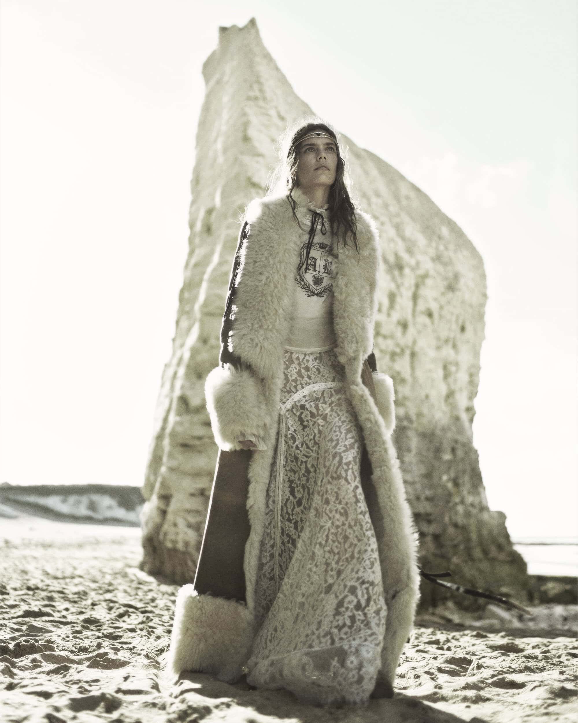 Model Betsy Gaghan standing on a sandy beach wearing a shearling-trimmed coat and white lace skirt against a massive chalk cliff.