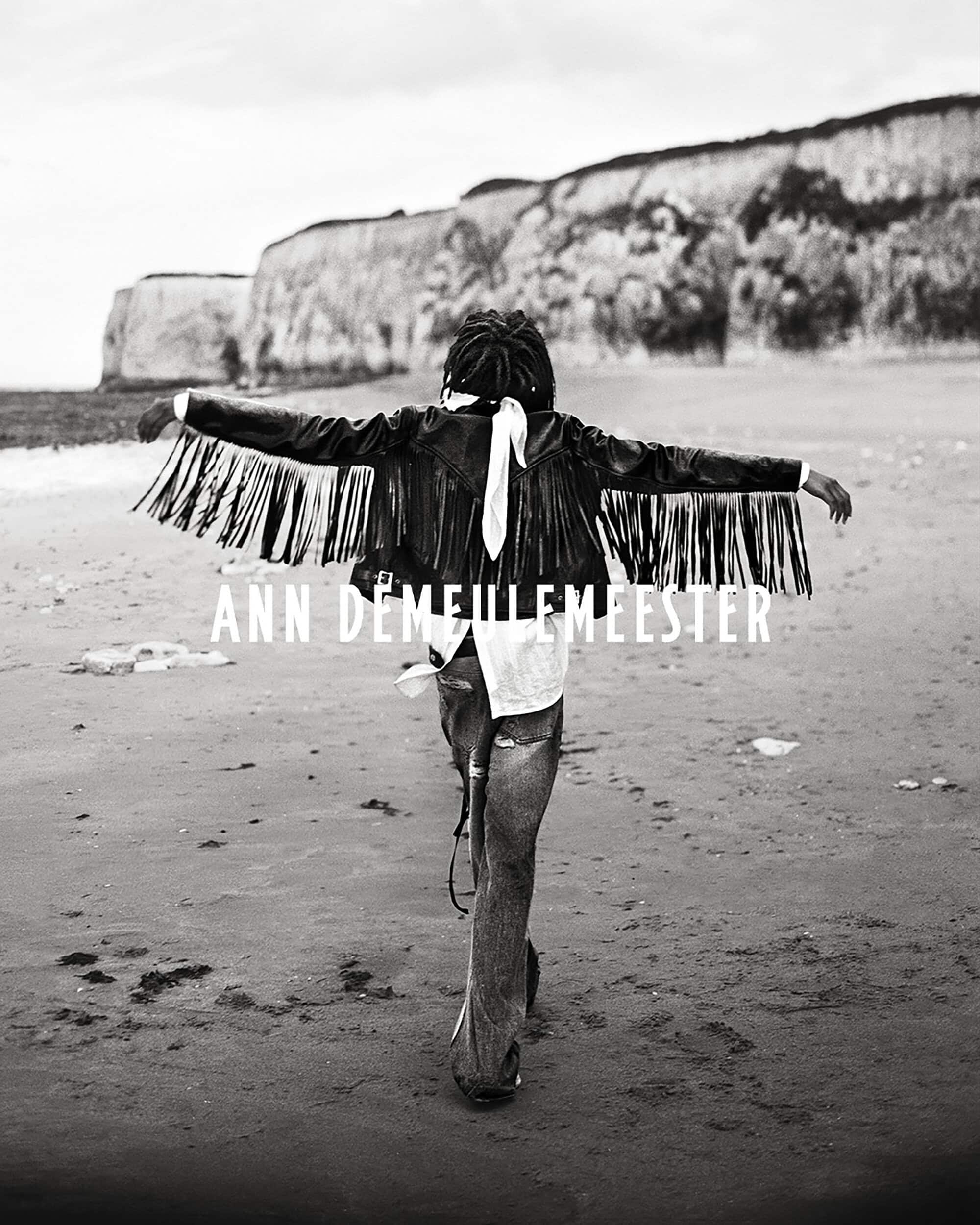 Model with arms outstretched wearing a black fringed leather jacket walking away on a sandy beach toward coastal cliffs.