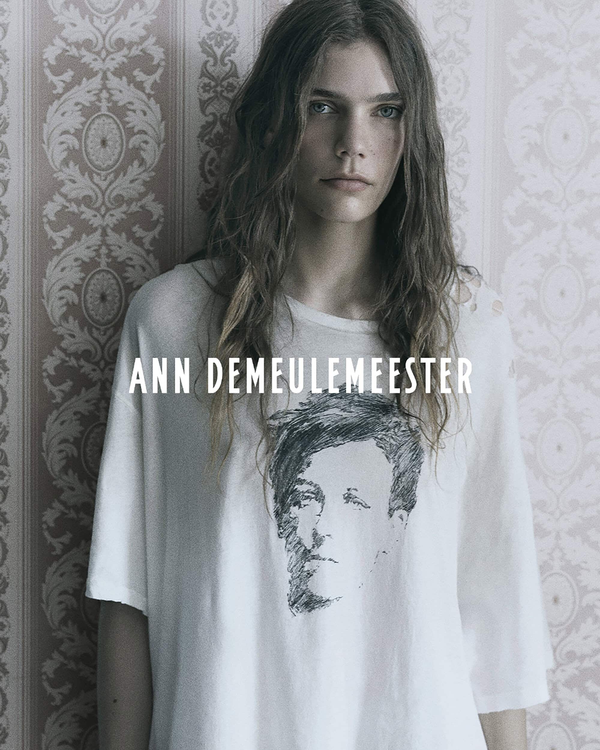 Model wearing an oversized white graphic t-shirt against patterned damask wallpaper in an Ann Demeulemeester campaign.