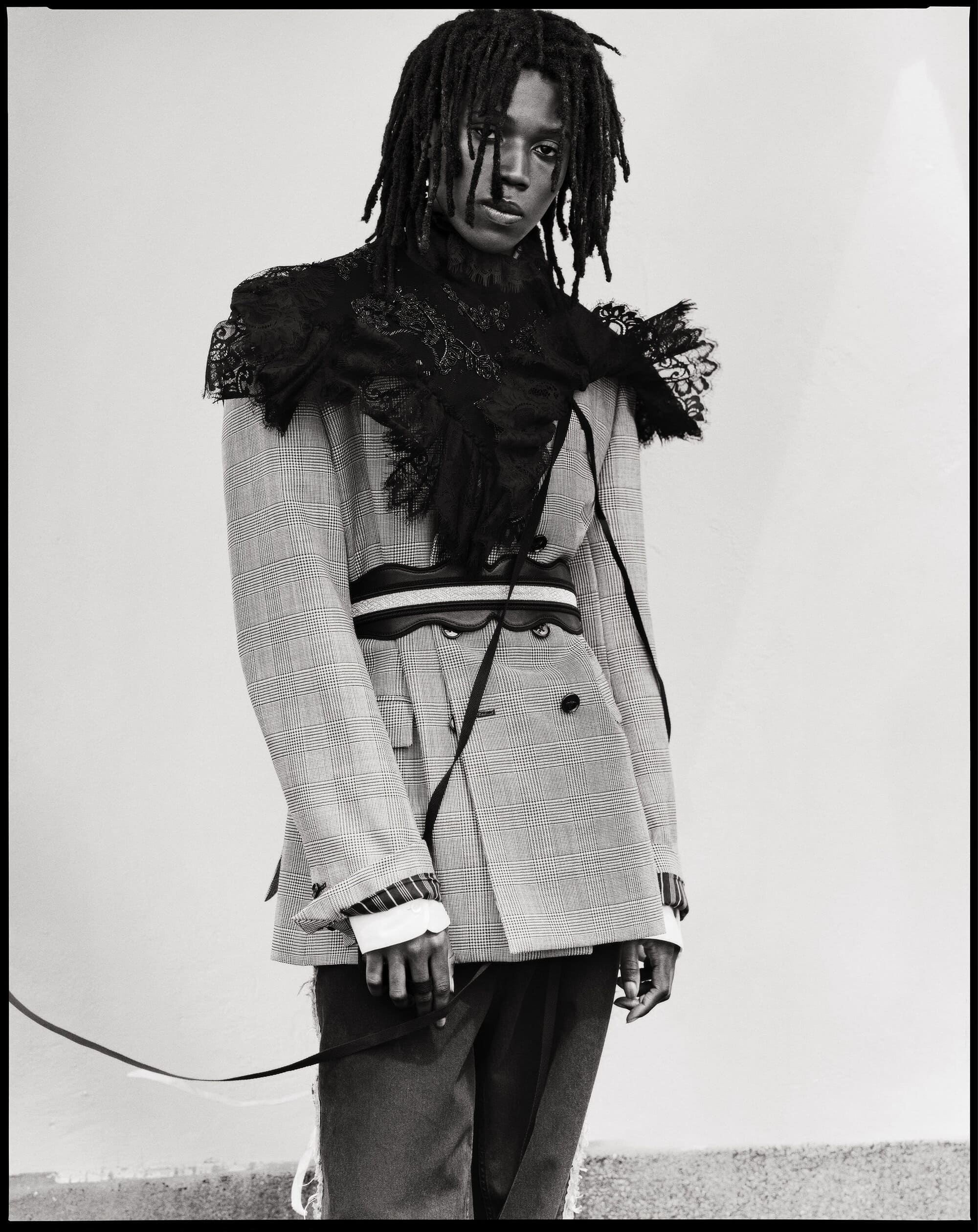 Model Ezekiel Conteh wearing a light tailored blazer with a black lace shoulder overlay and belt for Ann Demeulemeester.