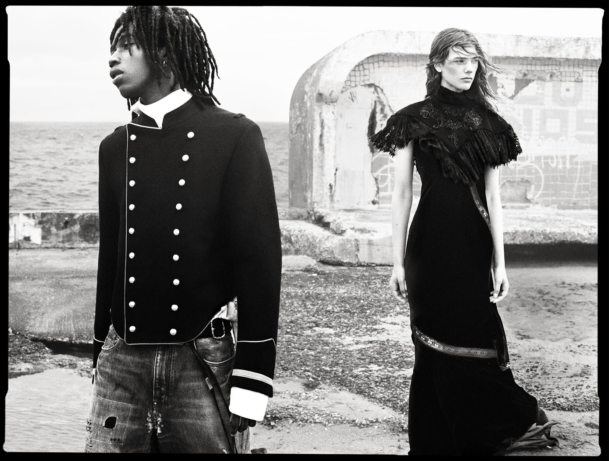 Models Ezekiel Conteh in a military jacket and Betsy Gaghan in a black ruffled dress outdoors by the coast in black and white.
