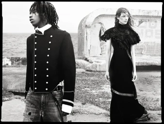 Models Ezekiel Conteh in a military jacket and Betsy Gaghan in a black ruffled dress outdoors by the coast in black and white.