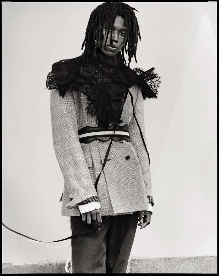 Model Ezekiel Conteh wearing a light tailored blazer with a black lace shoulder overlay and belt for Ann Demeulemeester.
