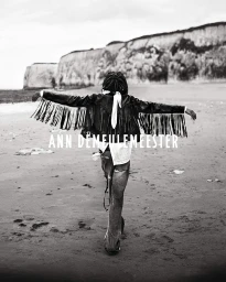 Model seen from behind with arms outstretched on a sandy beach wearing a black fringe jacket, with coastal cliffs in the distance.