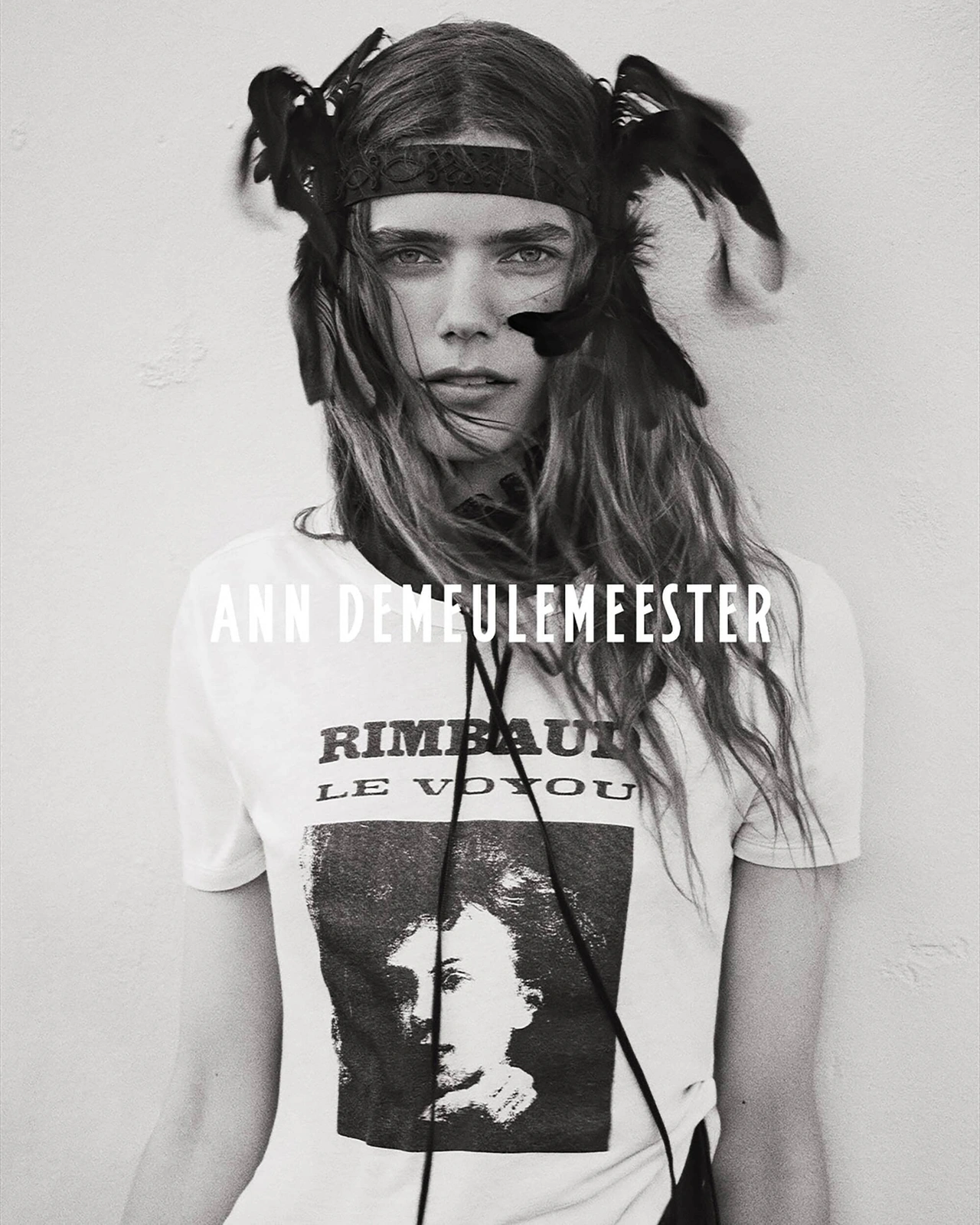 Model wearing a feathered headpiece and Rimbaud graphic t-shirt for Ann Demeulemeester against a plain light wall.