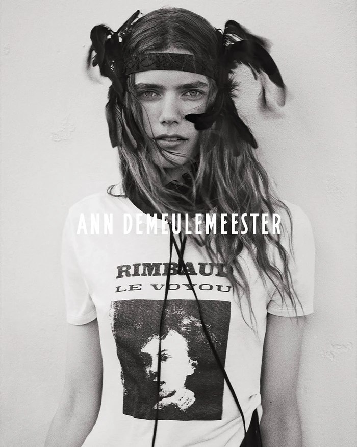 Model wearing a feathered headpiece and Rimbaud graphic t-shirt for Ann Demeulemeester against a plain light wall.