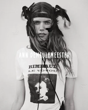 Model wearing a feathered headpiece and Rimbaud graphic t-shirt for Ann Demeulemeester against a plain light wall.