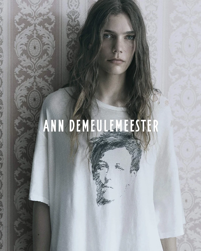 Model wearing an oversized white graphic t-shirt against patterned damask wallpaper in an Ann Demeulemeester campaign.