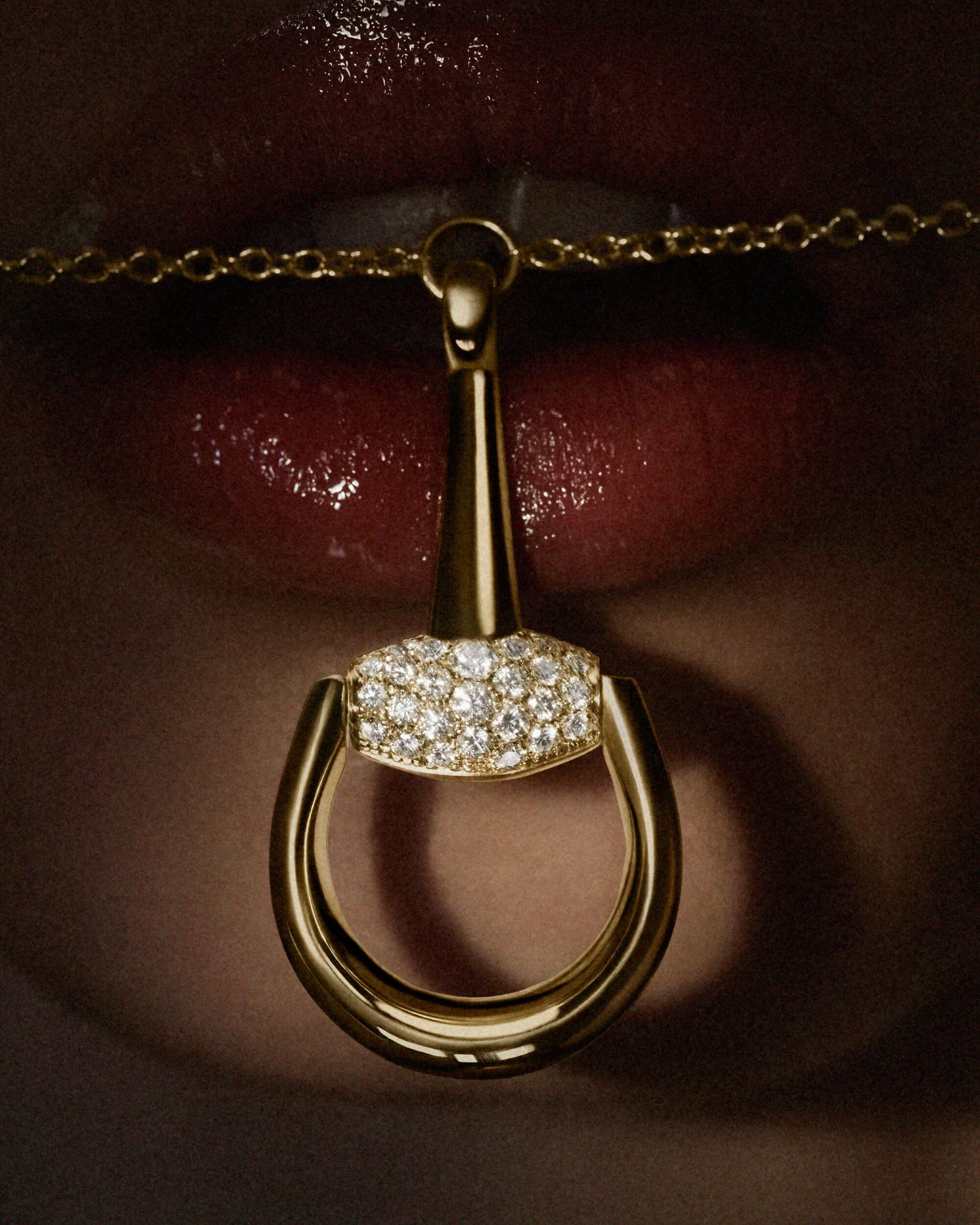 Close-up of a Gucci gold and pavé diamond Horsebit pendant necklace hanging in front of glossy crimson red lips.