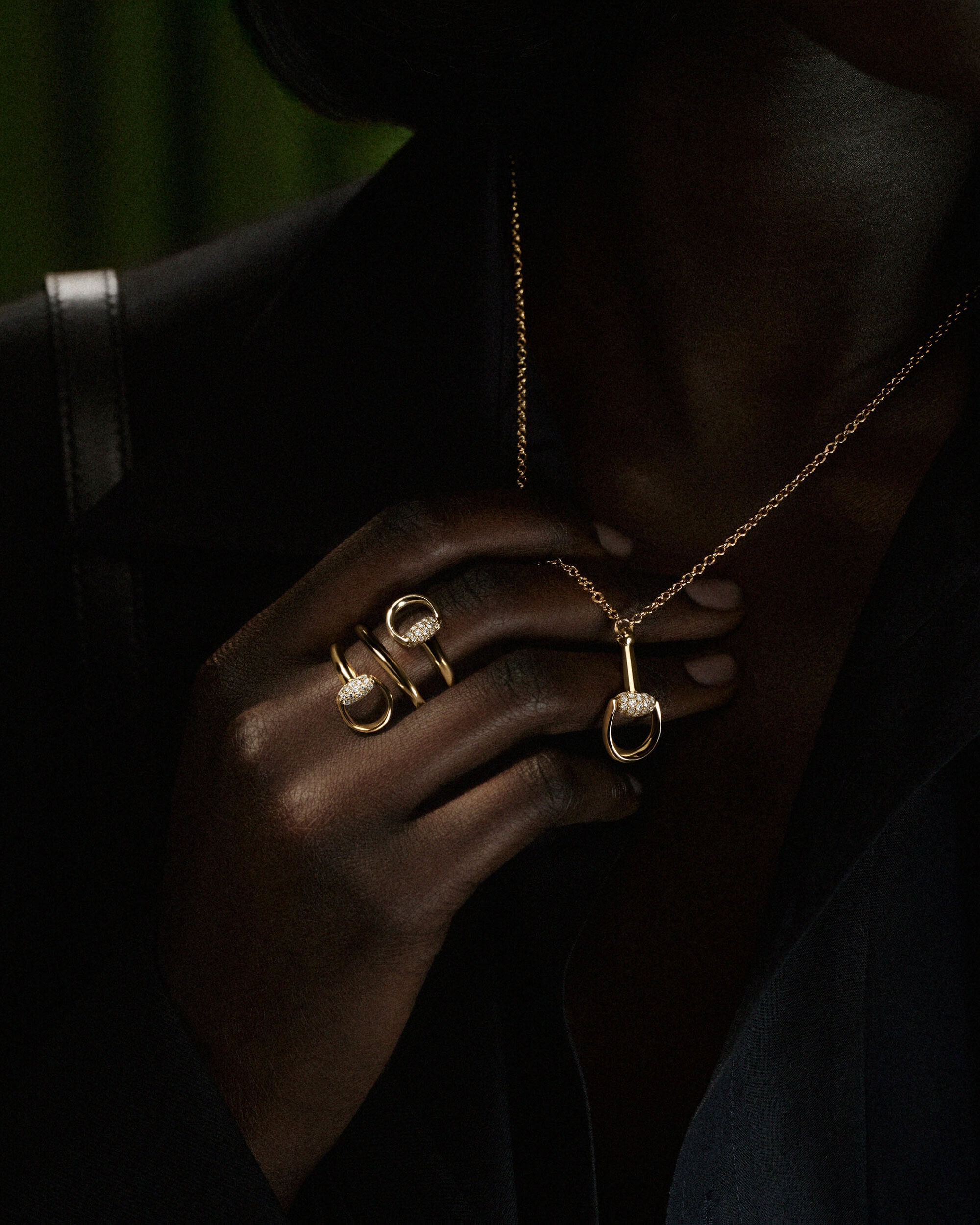Close-up of a person with dark skin wearing a gold horsebit necklace and coiled ring over a dark blazer.