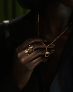 Close-up of a person with dark skin wearing a gold horsebit necklace and coiled ring over a dark blazer.