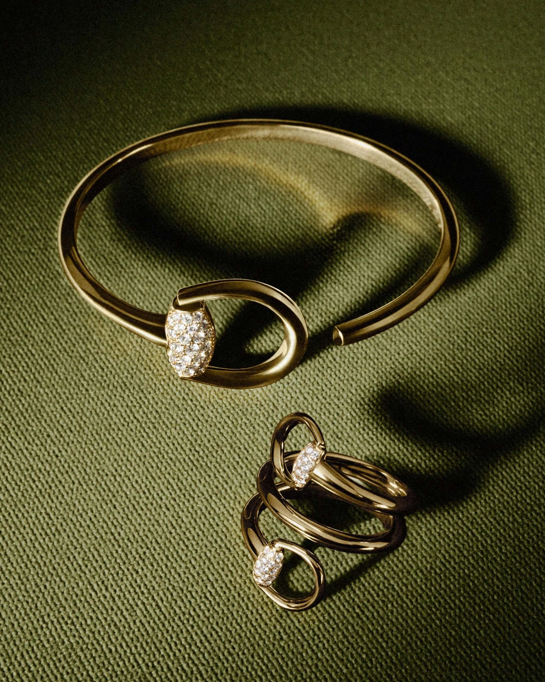 Gucci gold Horsebit cuff bracelet and matching spiral diamond ring displayed on olive green textured fabric.