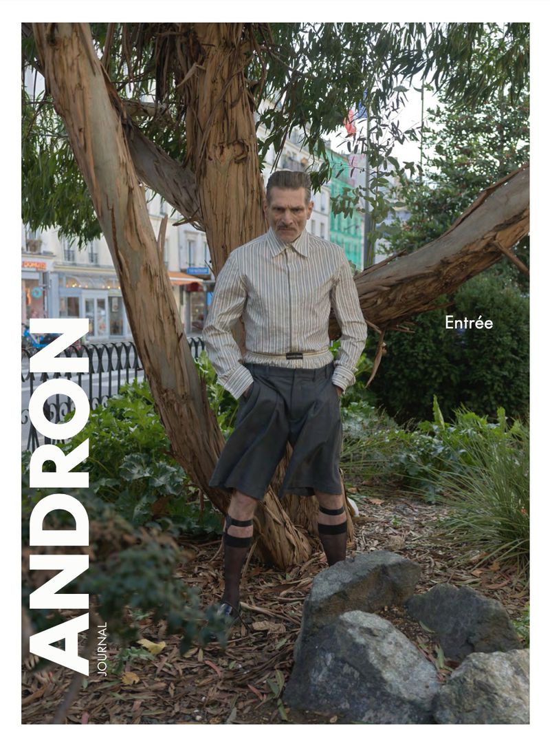 A man poses in a striped shirt and shorts in an urban park setting, alongside a tree and rock formation, with a building in the background.
