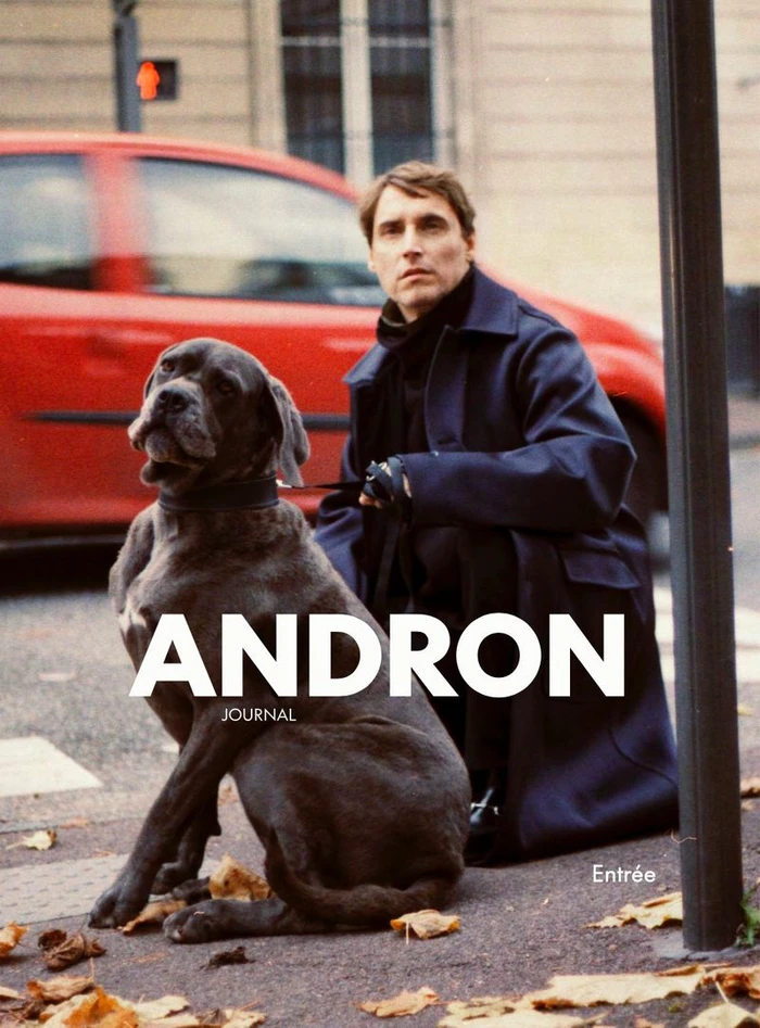 Man wearing a navy coat kneeling beside a large grey dog on a city street with a red car in the background