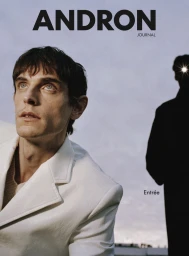 Man wearing a white blazer looking upward against a pale sky with a silhouetted figure in the background