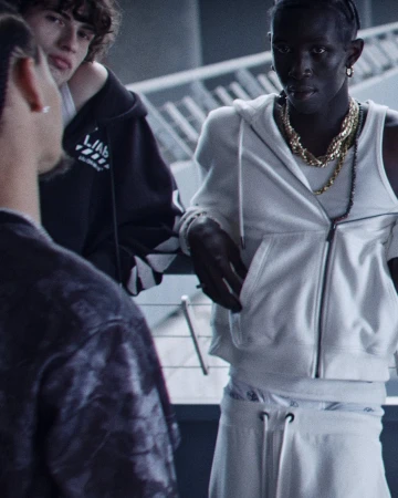 Black model wearing a white zip hoodie and layered gold chain necklaces standing alongside other models in an architectural setting.