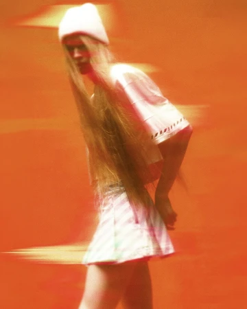 Model with long hair in a white beanie, t-shirt, and pleated skirt captured with motion blur against a bright orange backdrop.