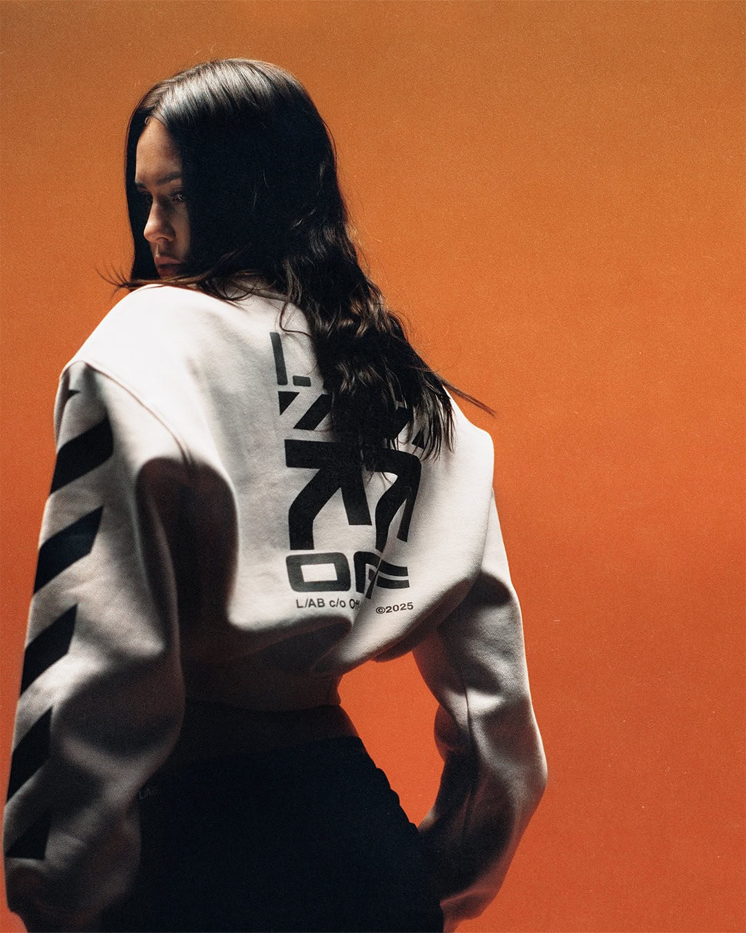 Model with long dark hair wearing a white cropped graphic sweatshirt and black pants against a vibrant orange studio backdrop.