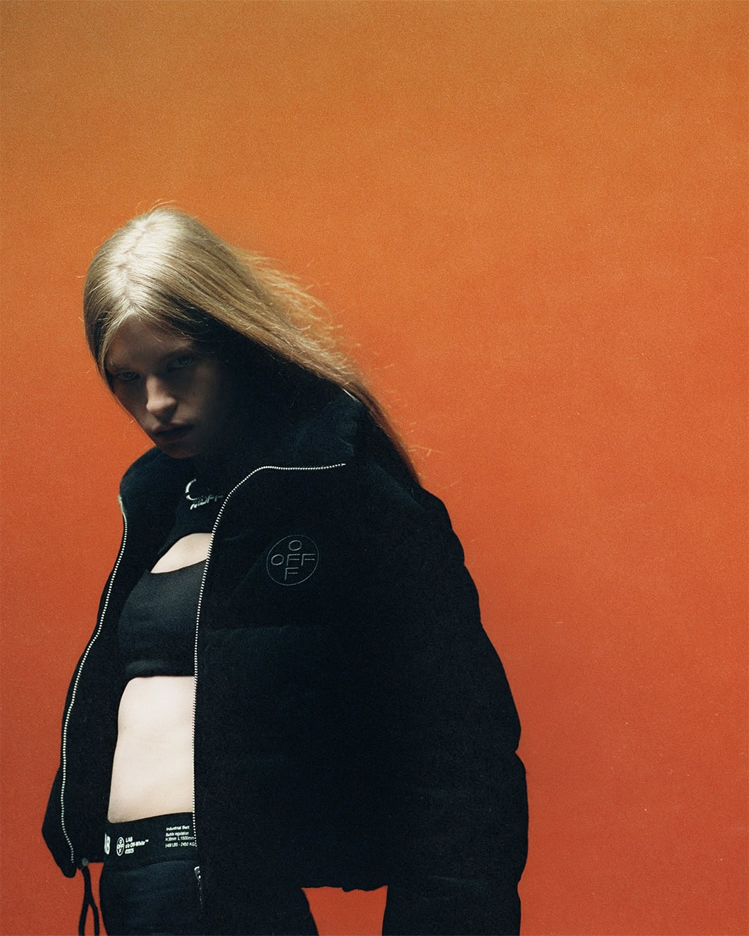 Model with blonde hair wearing a black puffer jacket and crop top against a vibrant orange backdrop.