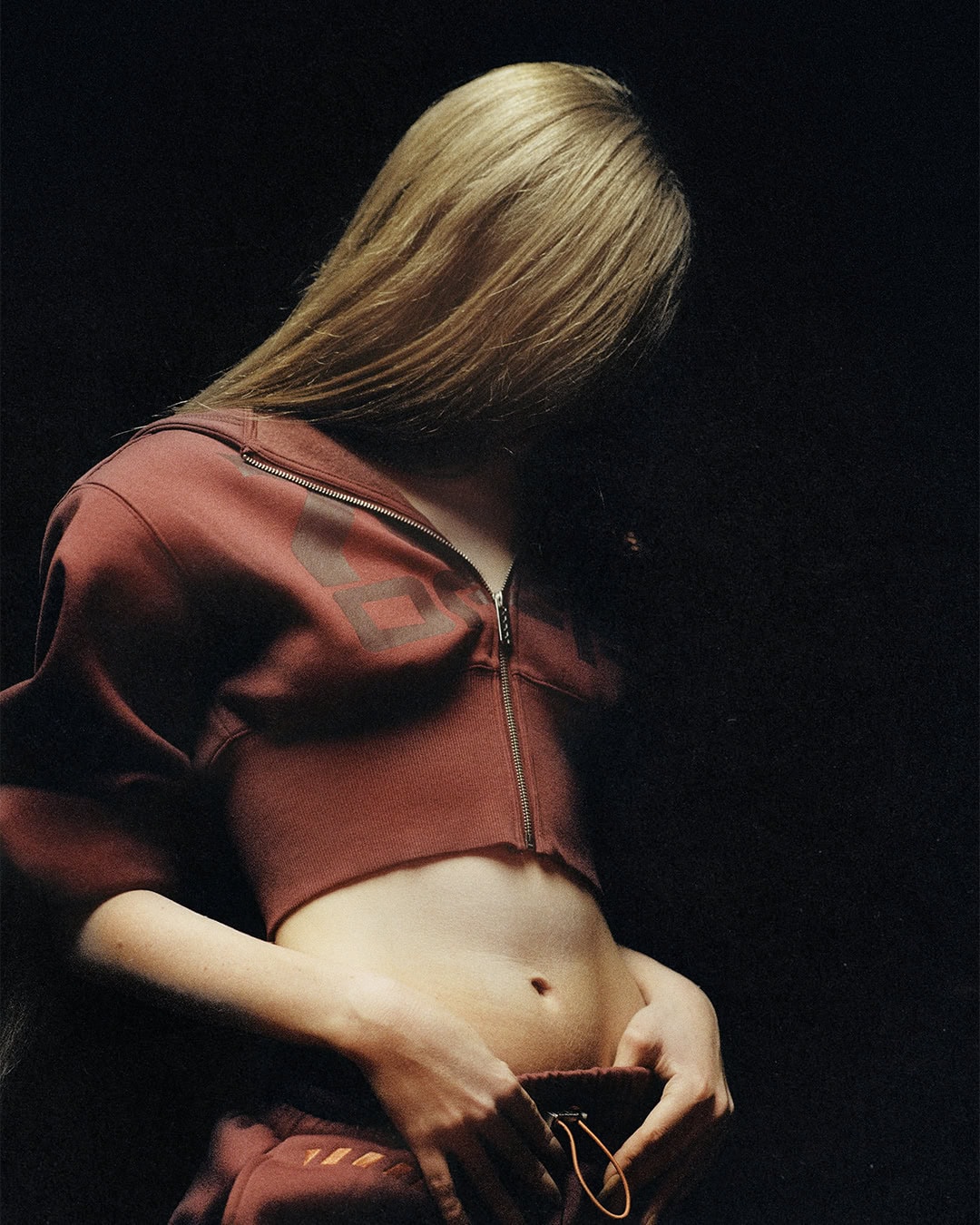 Model with long hair obscuring the face wearing a cropped maroon zip hoodie and low-rise sweatpants against a dark background.