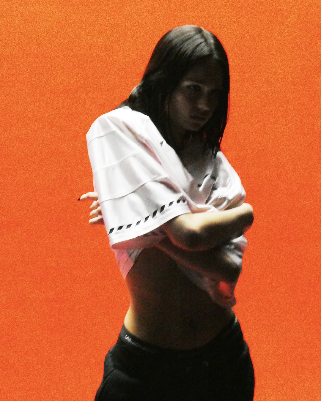 A model lifts a white graphic jersey to expose their midriff against a vibrant solid orange studio backdrop.