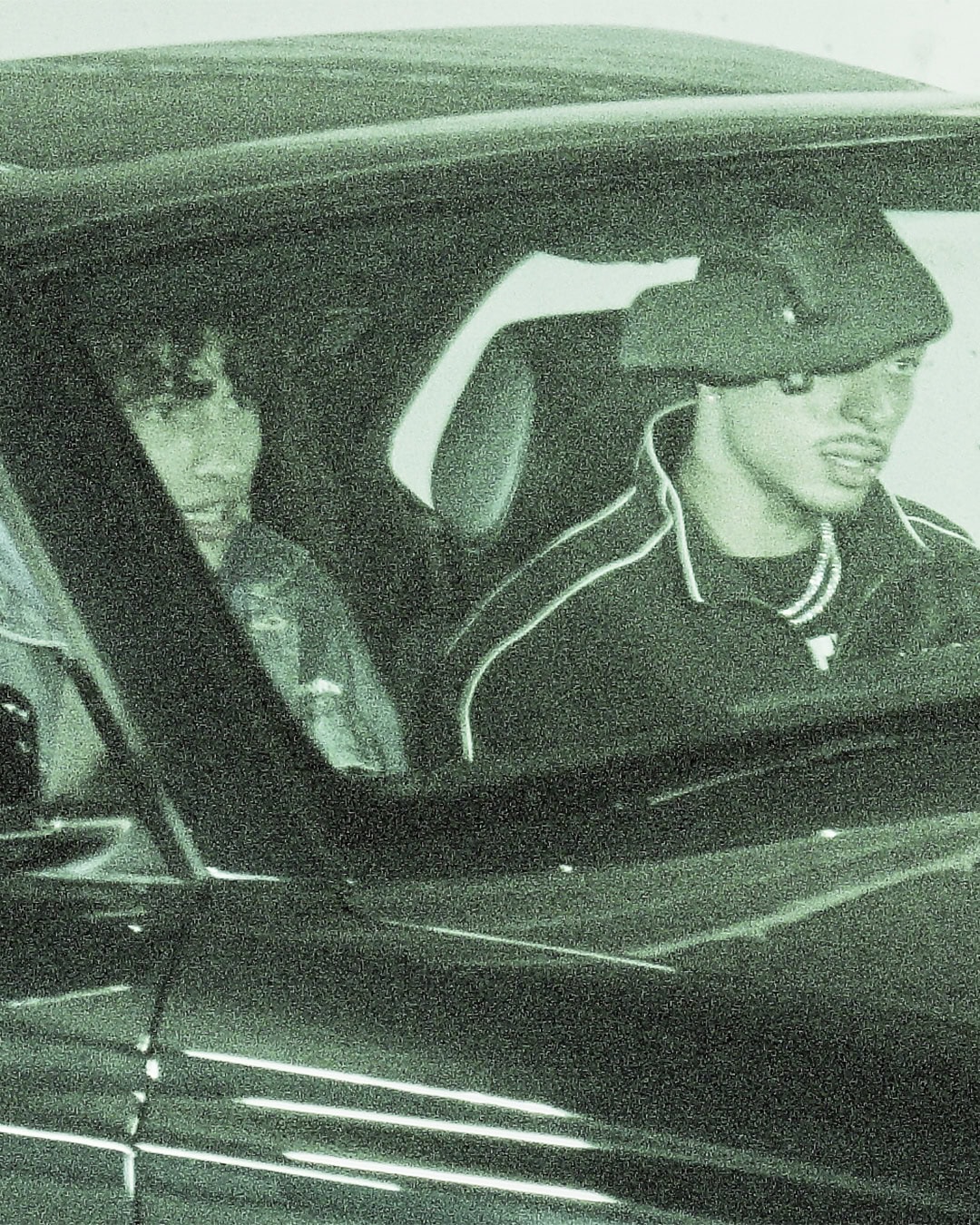 Two men seated inside a vehicle viewed through the front windshield in a green-tinted grainy surveillance aesthetic.