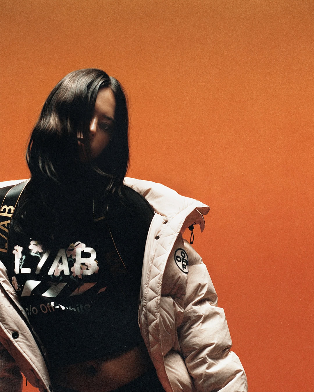 Model wearing a light beige puffer jacket over a black graphic crop top against a solid orange background.