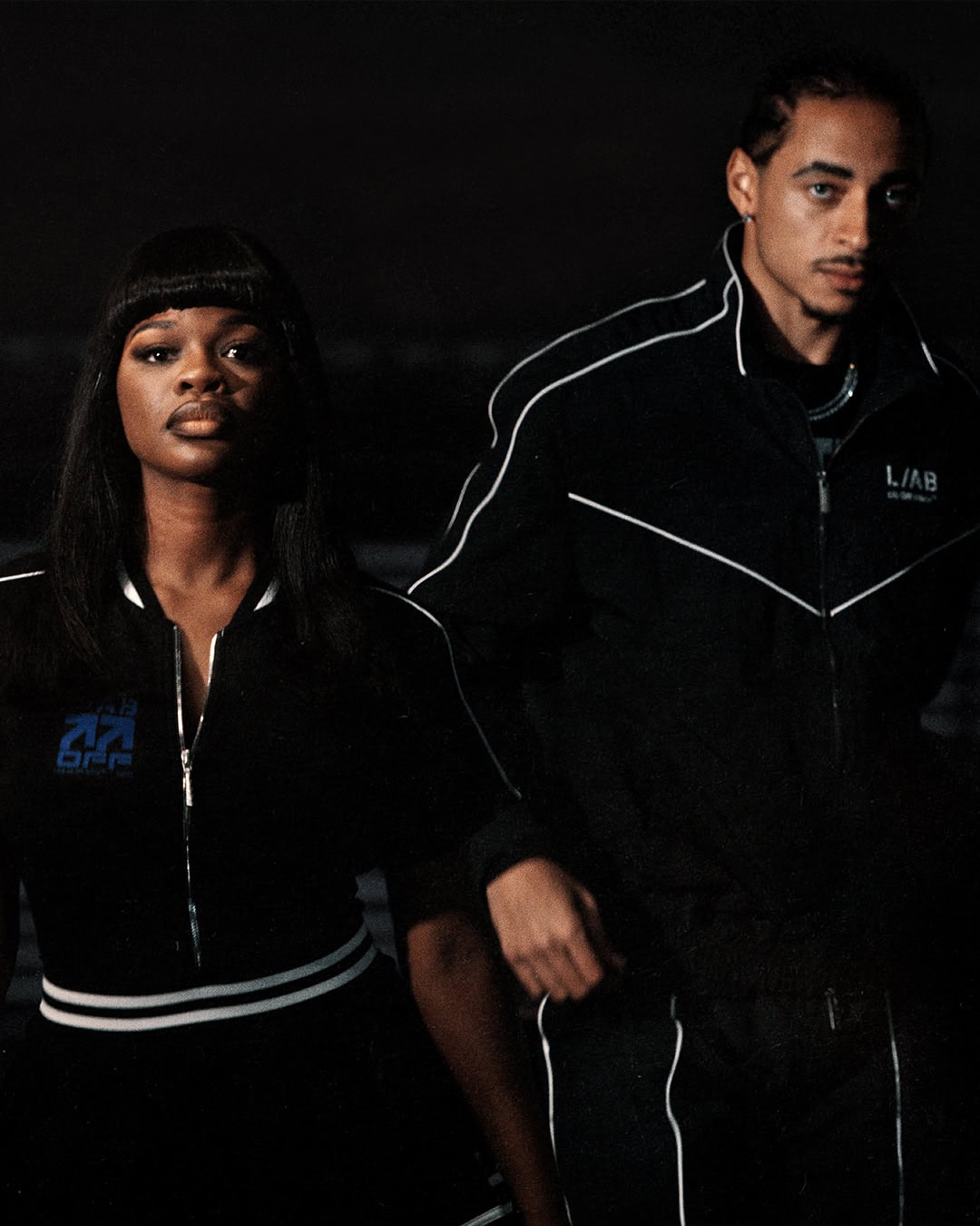 JT and male model posing in black tracksuits with white piping against a dark background for L/AB Off-White.