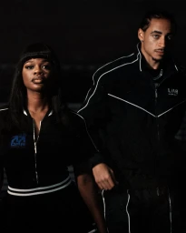 JT and male model posing in black tracksuits with white piping against a dark background for L/AB Off-White.