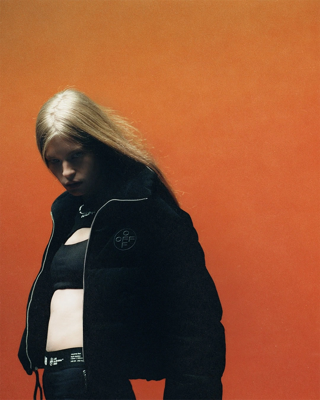 Model with blonde hair wearing a black puffer jacket and crop top against a vibrant orange backdrop.