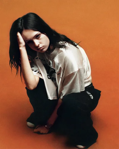 Model crouching in an oversized white jersey t-shirt and wide-leg black trousers against a solid warm orange studio background.