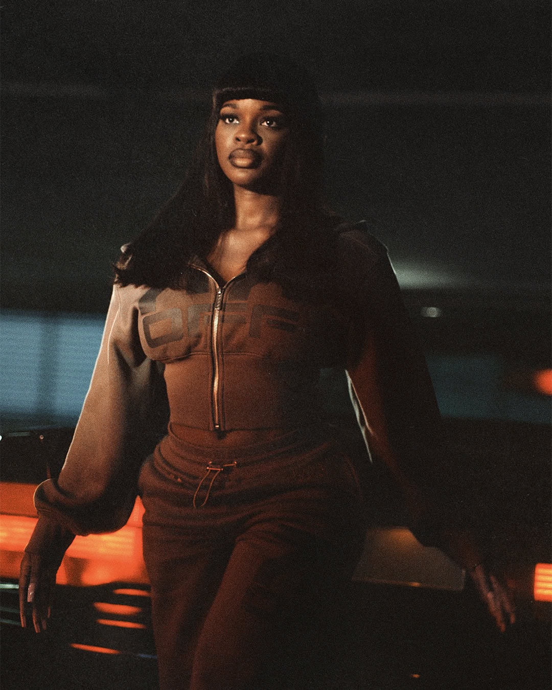 Model JT posing in a brown cropped zip-up tracksuit in front of illuminated glowing car lights in a dark garage setting.