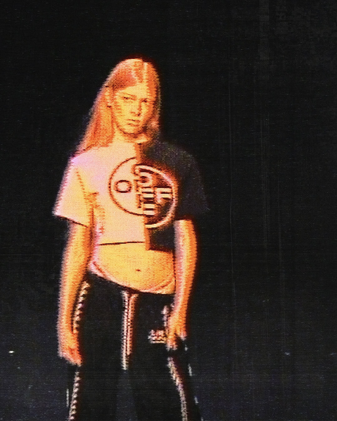 Model wearing a two-tone cropped graphic tee and black track pants against a dark background with lo-fi scan line texture.
