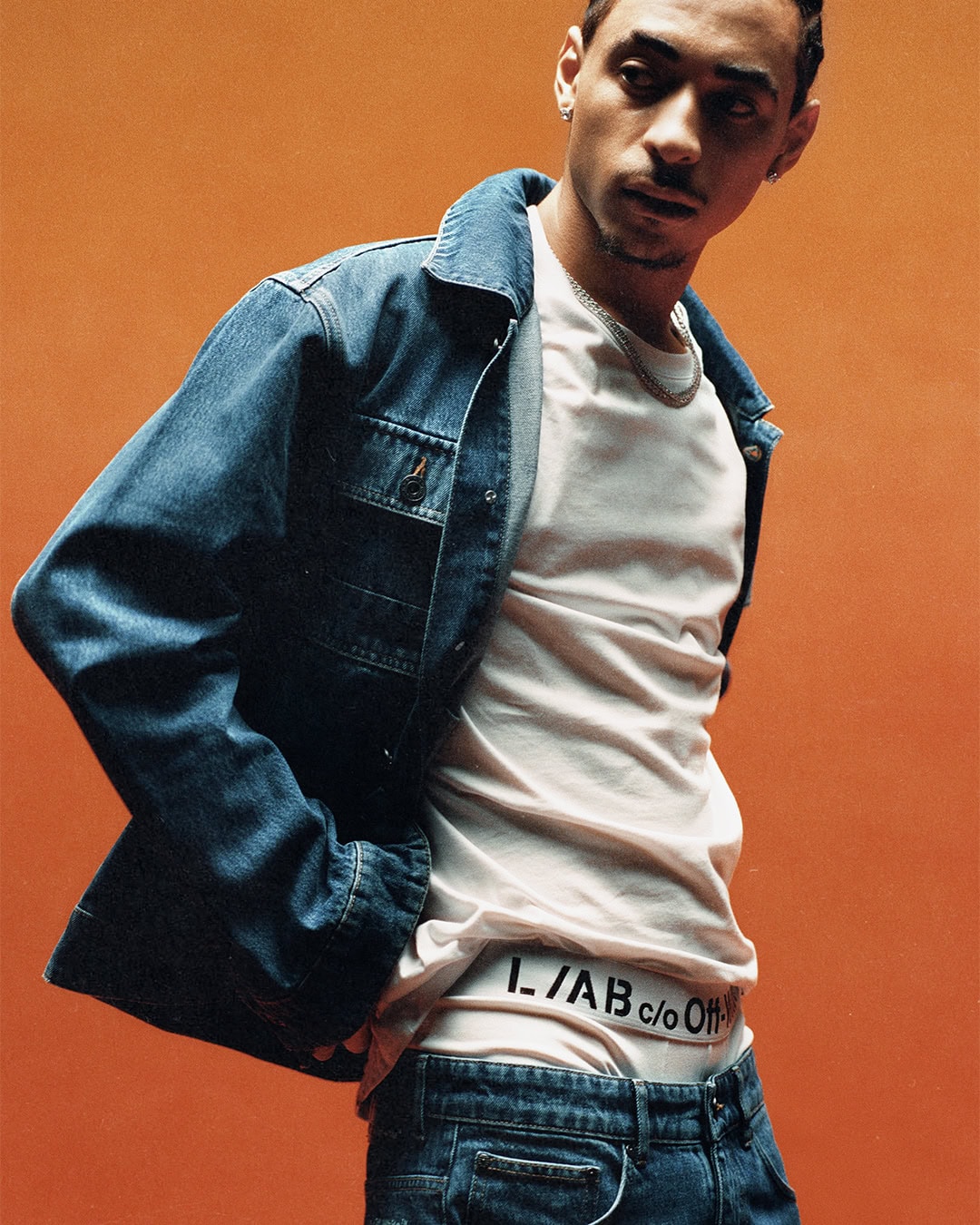 A man posing in a blue denim jacket, white t-shirt, and jeans showing an L/AB c/o Off-White waistband against an orange backdrop.
