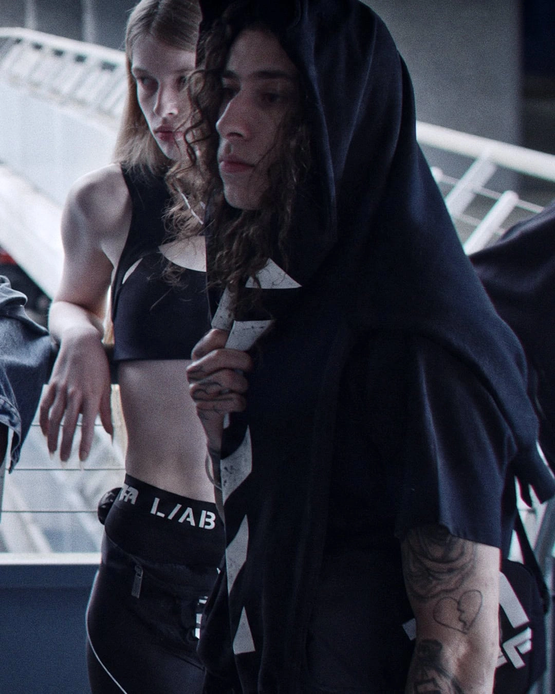 Models wearing black streetwear garments and branded L/AB leggings standing near an outdoor bridge railing.