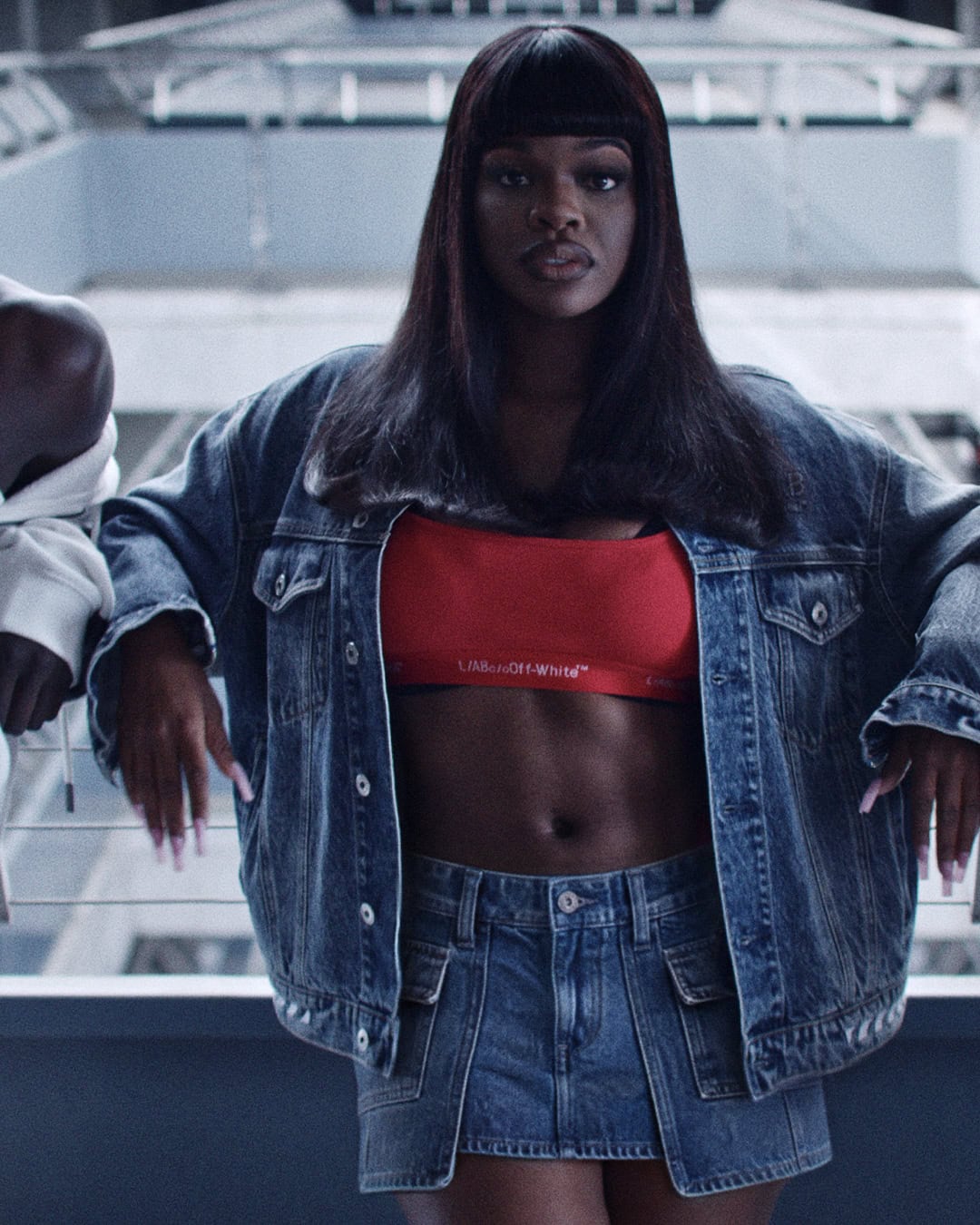 JT wearing an oversized denim jacket, red crop top, and denim skirt while leaning against a railing.