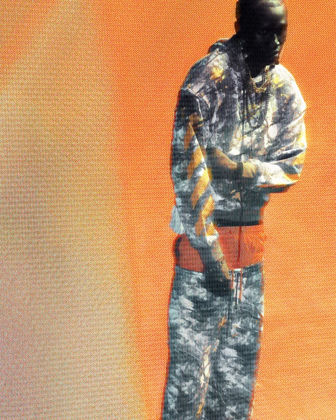 A Black male model stands in a printed grey and yellow hoodie with matching trousers against a bright orange studio backdrop.
