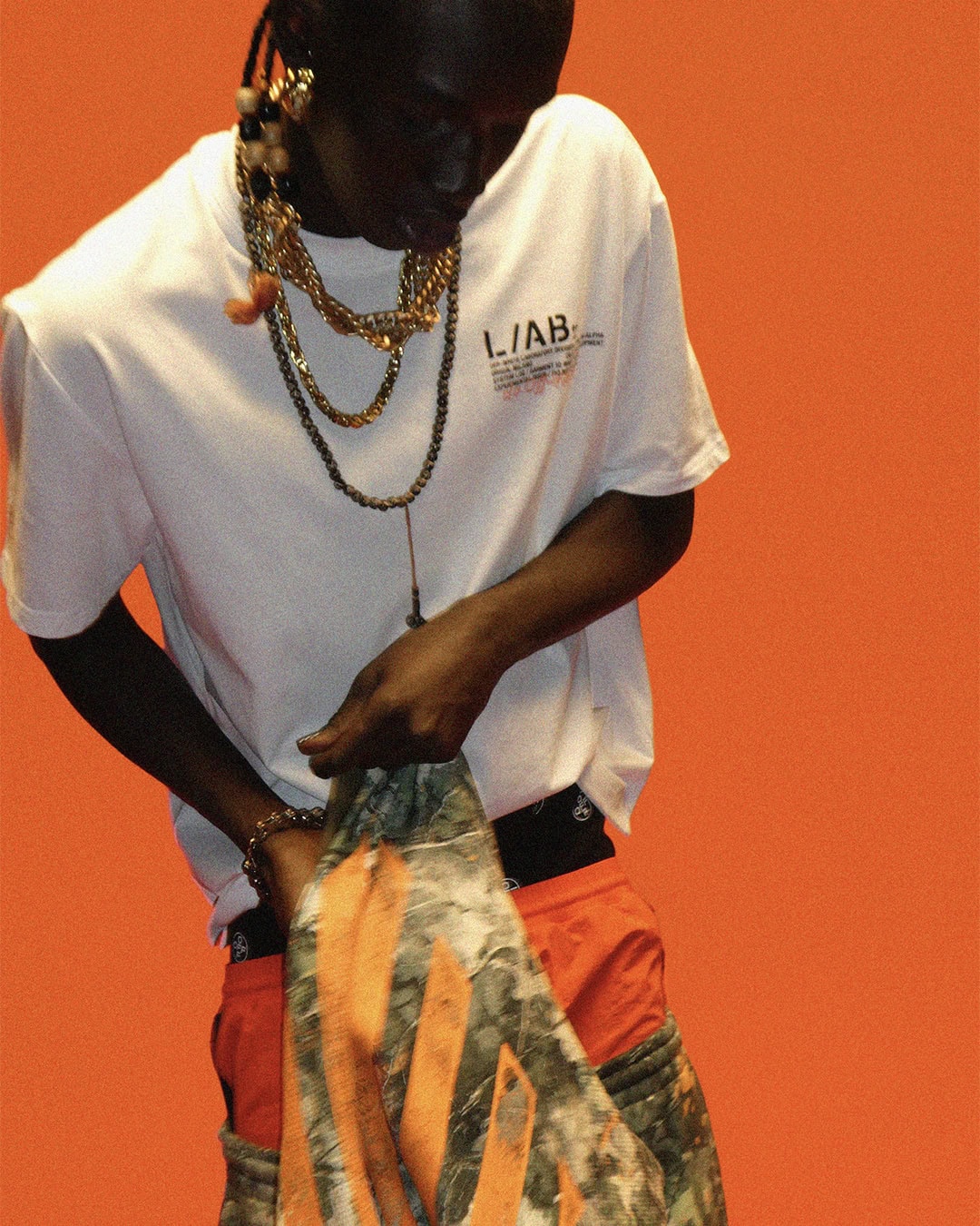 A Black man in a white L/AB Off-White graphic t-shirt and layered gold chain necklaces looks down against an orange studio backdrop.