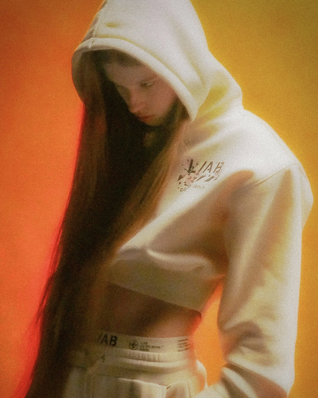 Model wearing a cream cropped hoodie and sweatpants with very long hair against an orange and yellow gradient backdrop.
